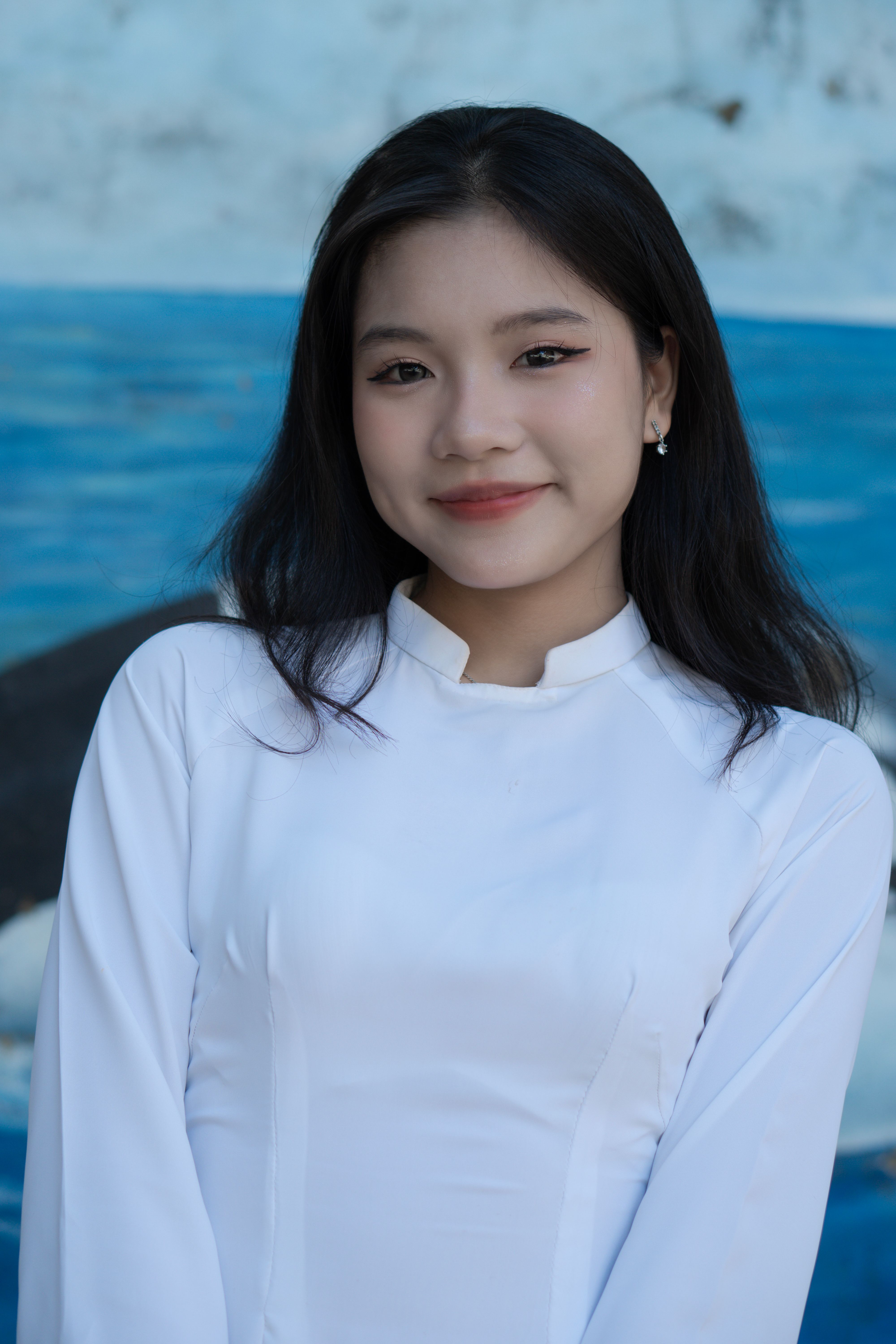 A young woman wears a traditional ao dai, smiling against a blue background outdoors.