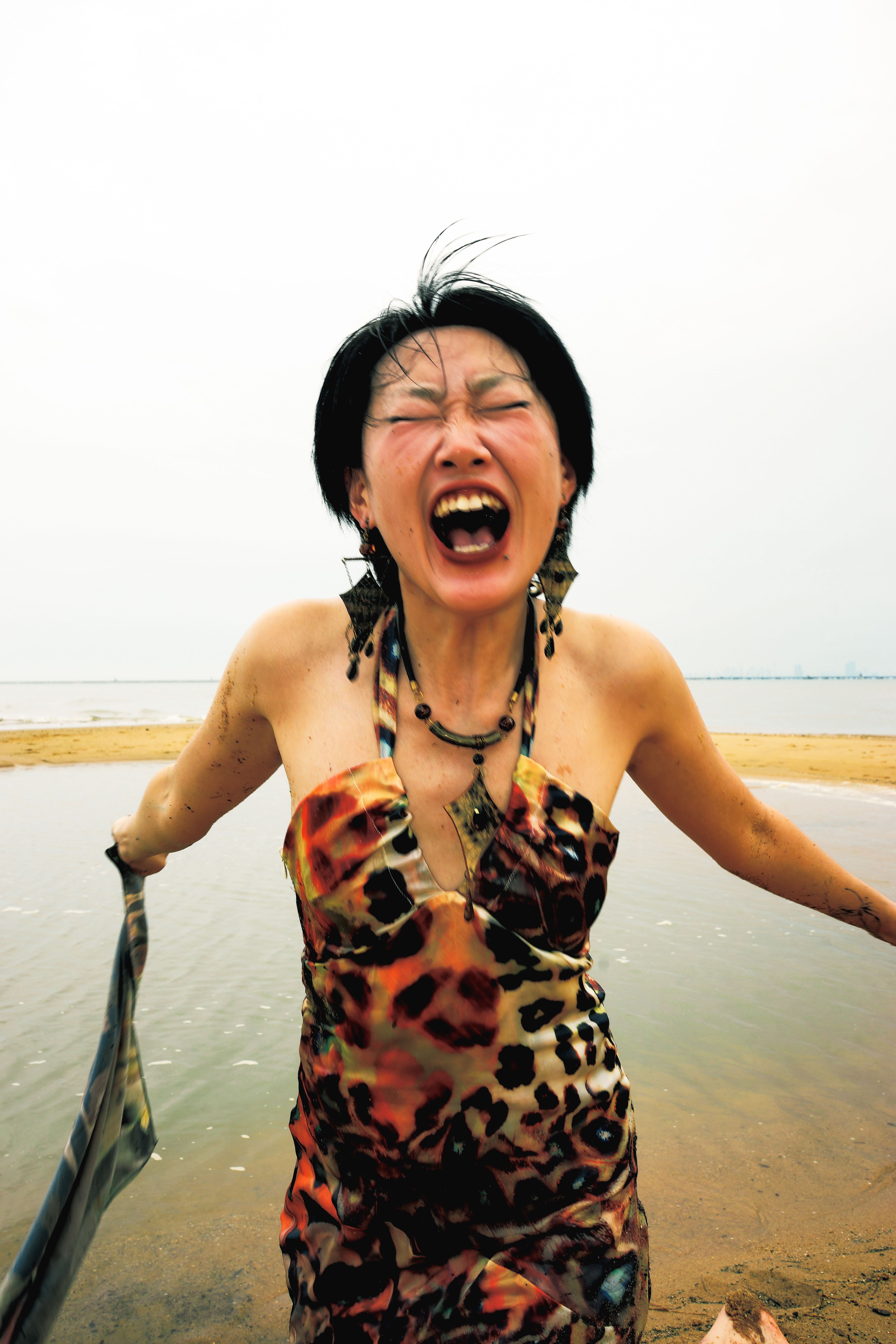 Asian woman with eyes closed joyfully at the beach in a colorful dress, expressing happiness and freedom.