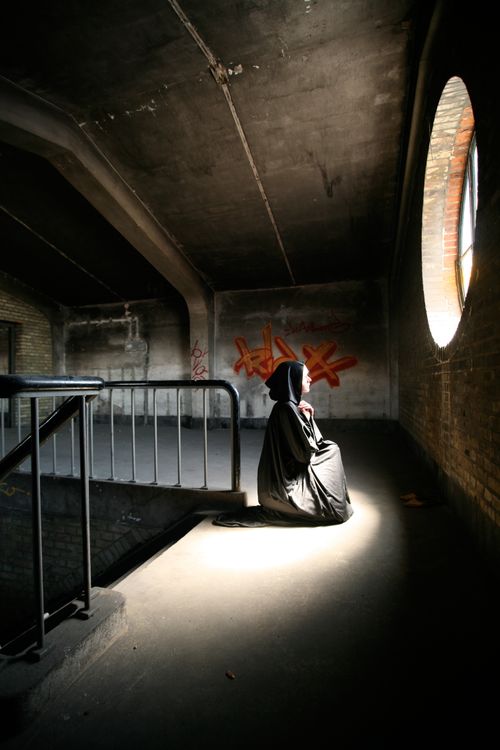 Free A solitary figure in a black cloak kneels in a dimly lit attic space, creating an eerie atmosphere. Stock Photo
