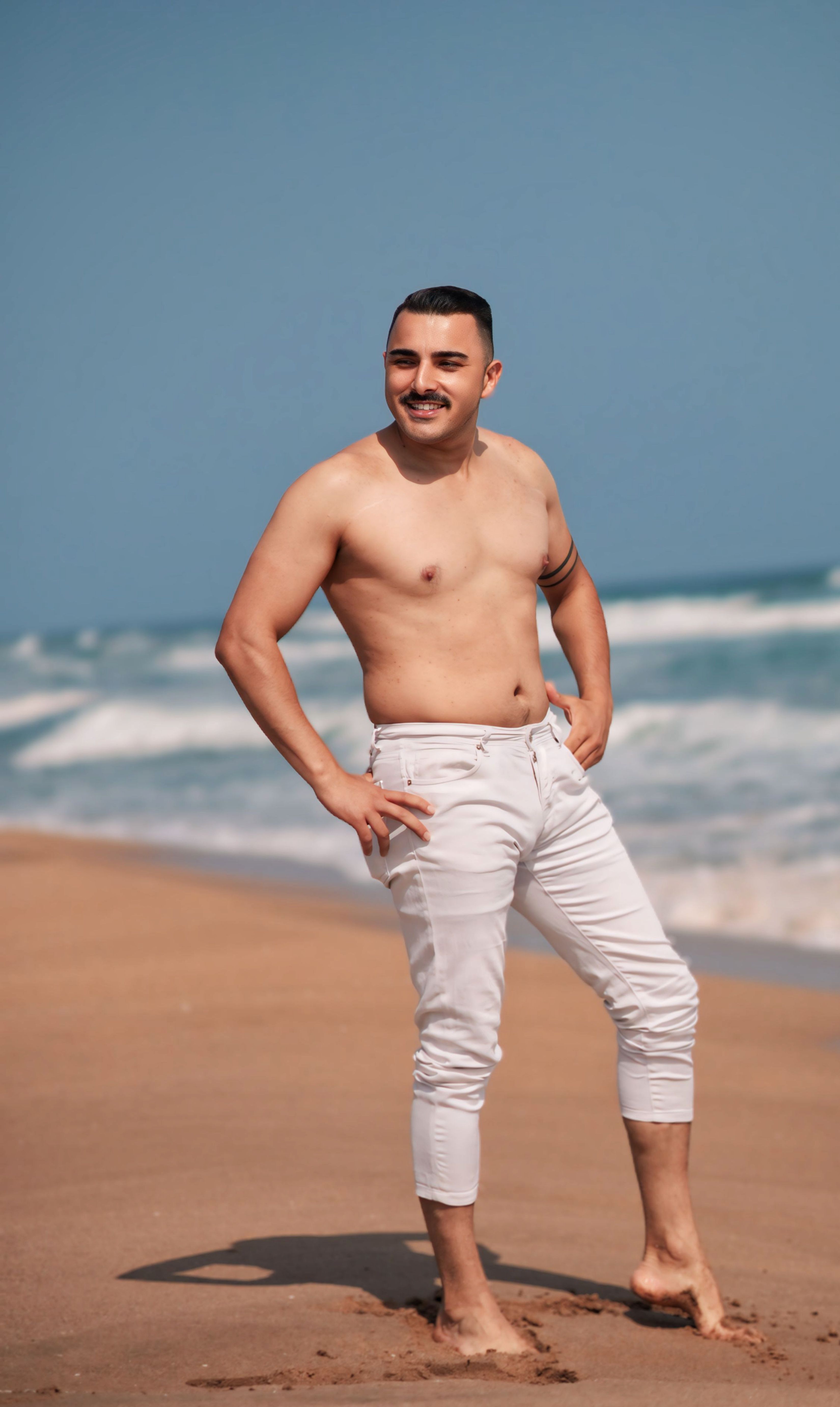 Confident man standing on the beach in white pants with the ocean in the background.