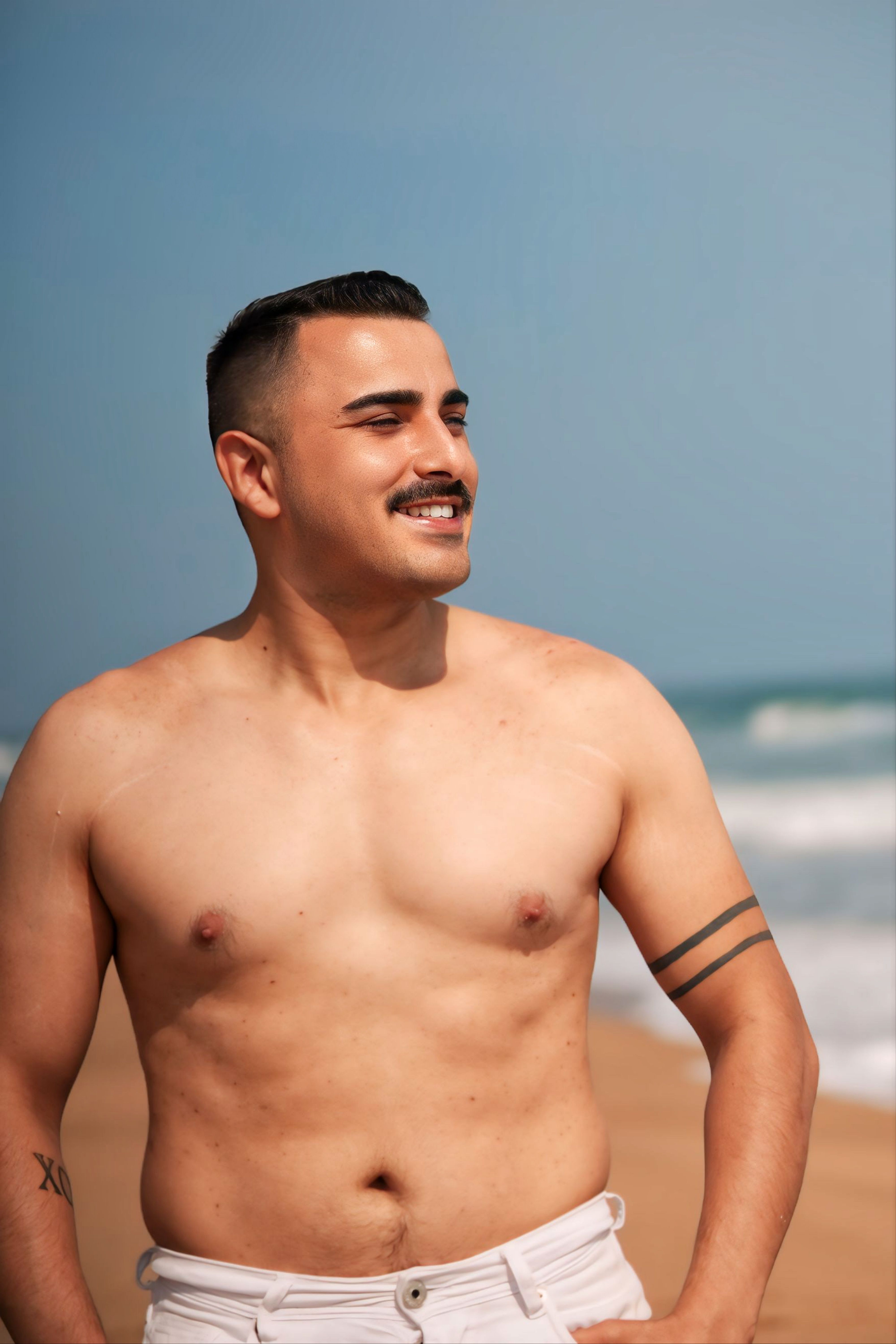 A shirtless, tattooed man with a mustache enjoys a sunny day at the beach, exuding happiness and relaxation.