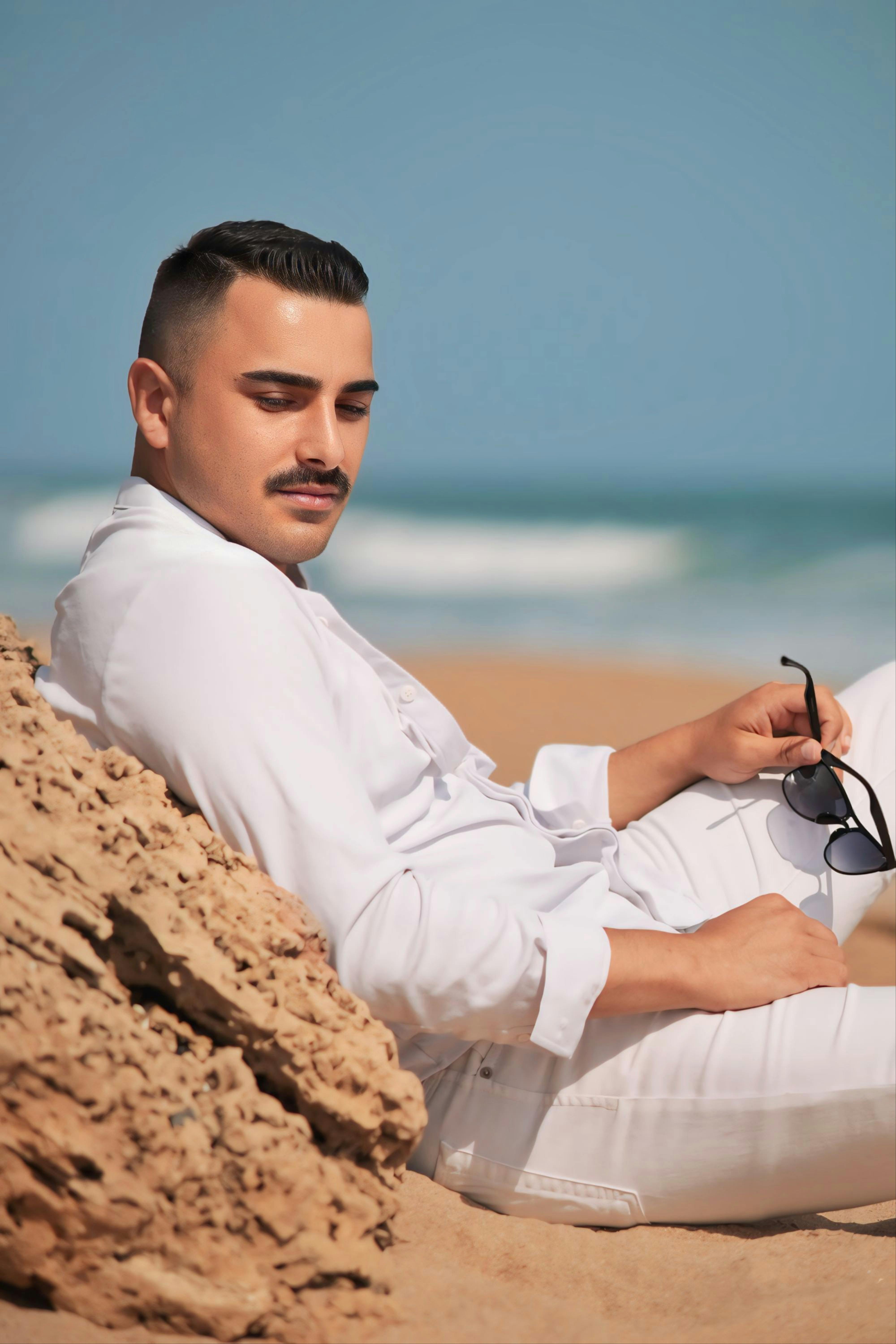 Fashionable man in white relaxing on a sunny beach, exuding calm and style.
