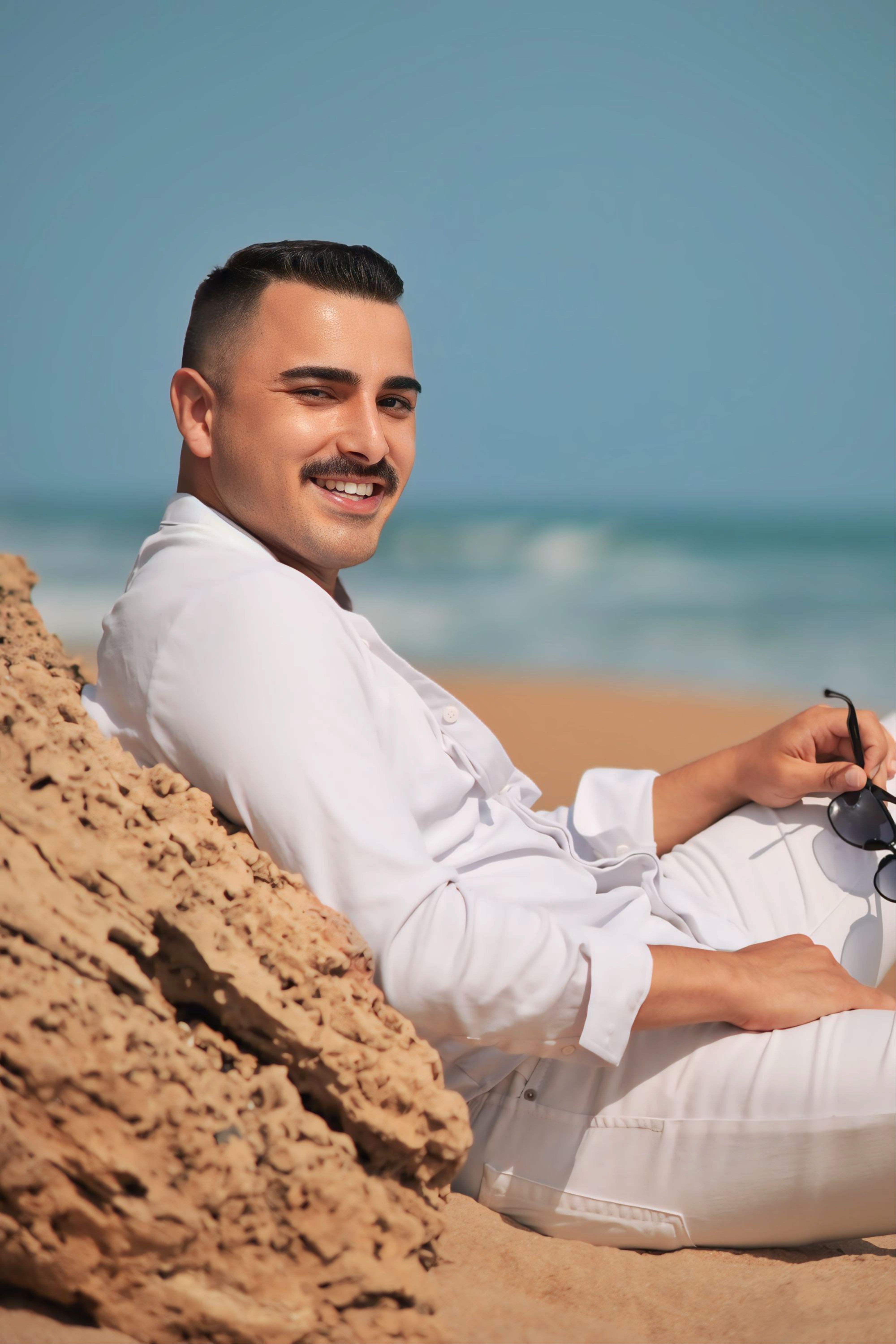 Smiling man in white clothes relaxing by the beach with clear blue sky.