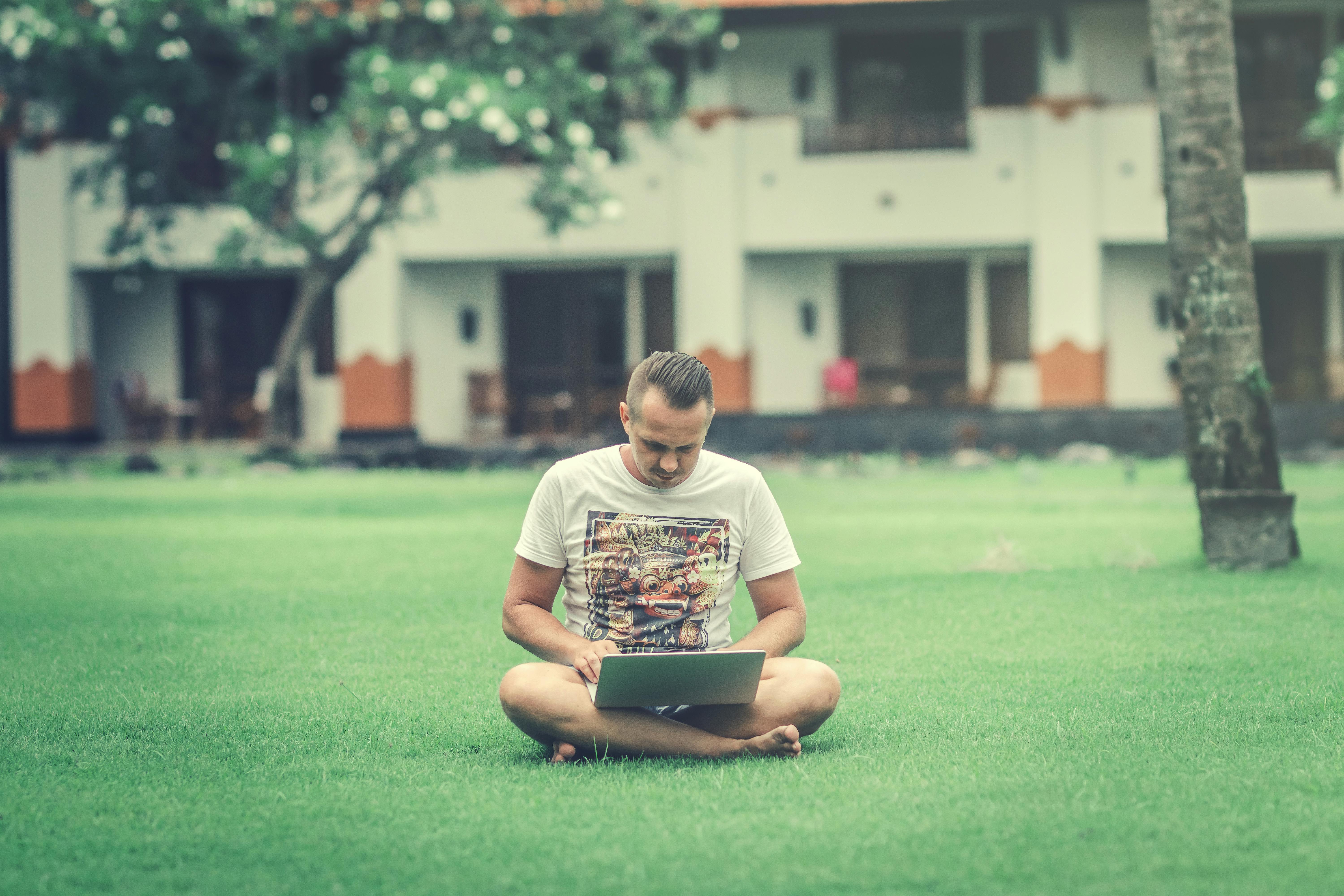 Photo of a Man in White Printed T-shirt Sitting on Green Grass in view of a Building Using a Laptop