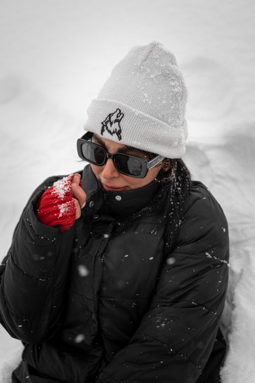 Free Woman in winter attire with sunglasses, black jacket, white hat, and red gloves in a snowy setting. Stock Photo