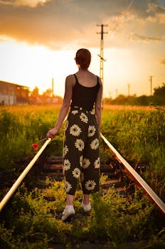 日落时分，一个女人手持一朵花走在铁轨上，营造出宁静的氛围