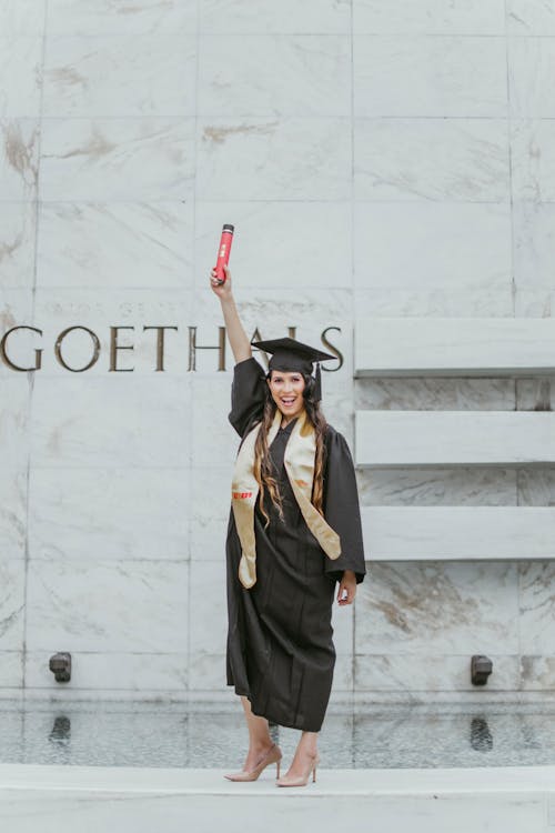 卒業証書を保持している大理石の壁の前に立っている黒いアカデミックドレスの笑顔の女性の写真