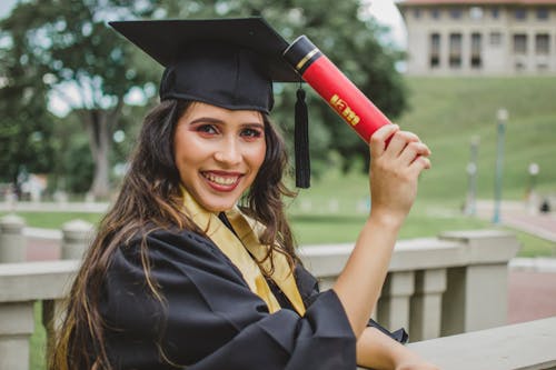 Foto De Retrato De Enfoque Selectivo De Mujer Sonriente En Vestido Negro Académico Sosteniendo Diploma Posando