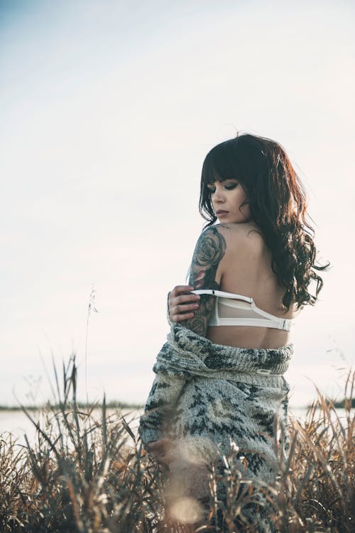 Free Photo of Tattooed Woman in Grey Sweater and White Bra Posing by Grass Next to Body of Water Stock Photo