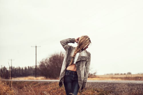 Photo of Woman in a Gray Sweater, White Tank Top, and Blue Jeans Standing on the Side of the Road Posing While Looking Away