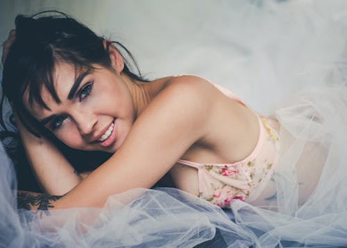 Free 白い薄手の布の上に横たわっているピンクの花のブラジャーを着ている笑顔の女性の写真を閉じる Stock Photo