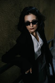 Stylish woman in black blazer and sunglasses poses confidently in a shadowy urban environment in Baku.