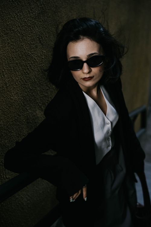 Free Stylish woman in black blazer and sunglasses poses confidently in a shadowy urban environment in Baku. Stock Photo