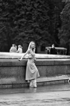 Stylish woman in dress leans against fountain in a city park, black and white.