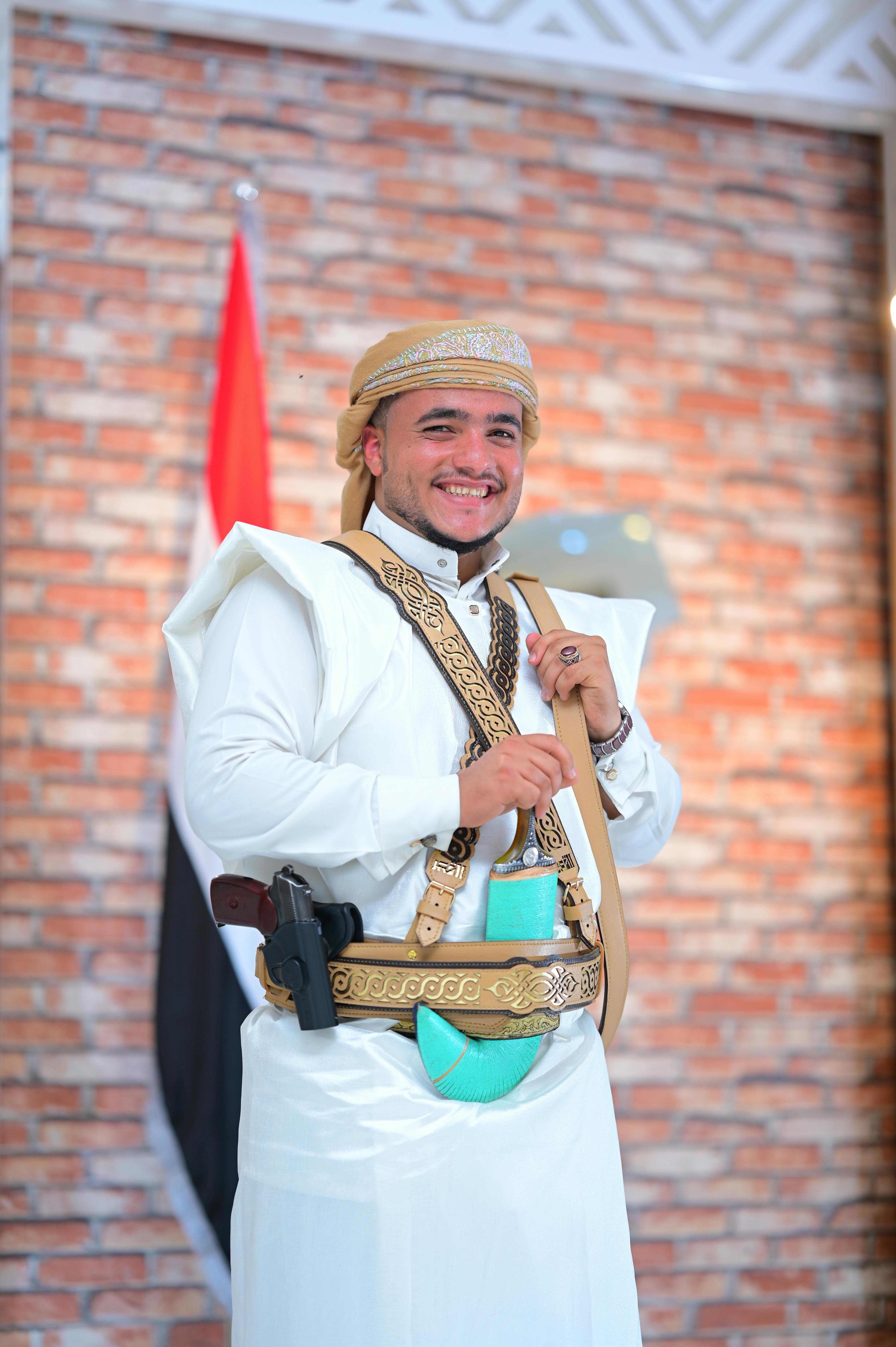 Man in traditional attire with ceremonial weapons and Yemen flag in background.