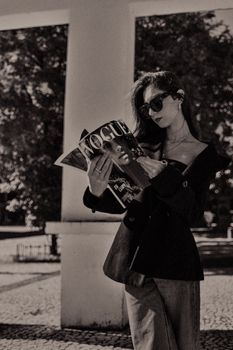 A fashionable woman wearing sunglasses reads Vogue magazine outside, exuding classic style.
