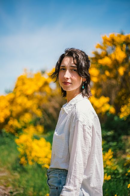 Portrait of a young woman outdoors surrounded by vibrant yellow flowers, under a clear blue sky.