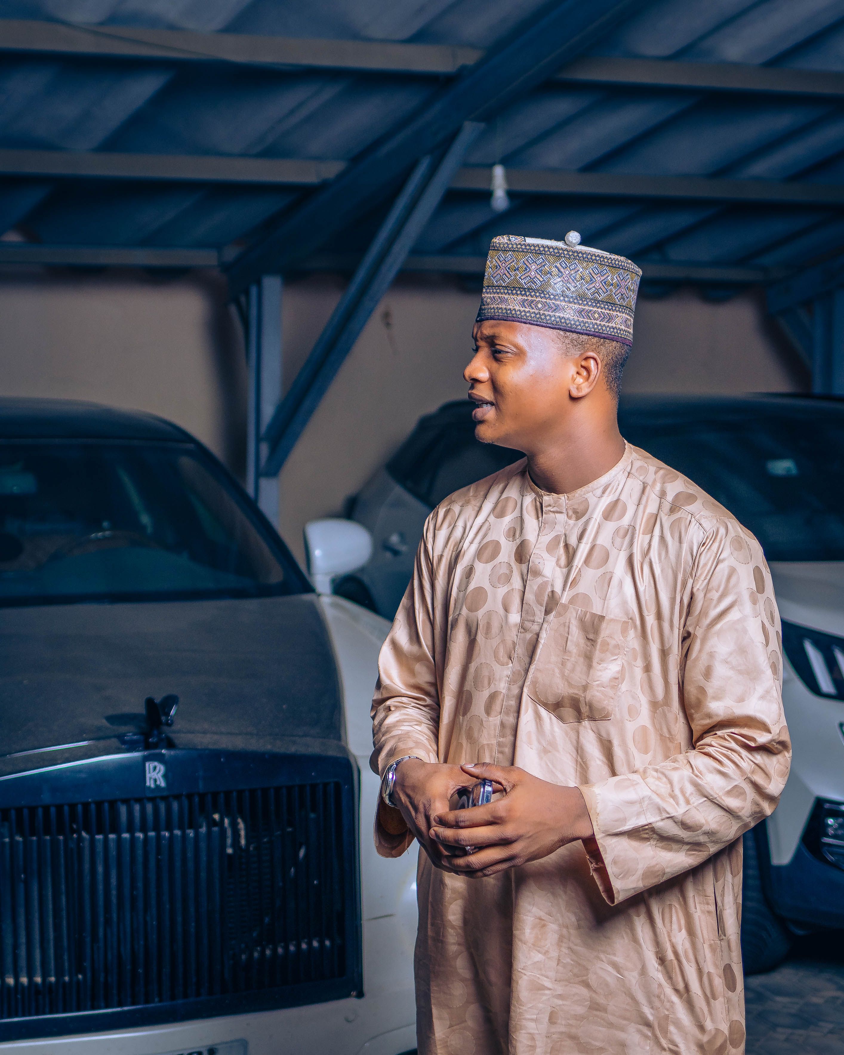 Man in traditional African attire standing next to a luxury car in a covered parking area.