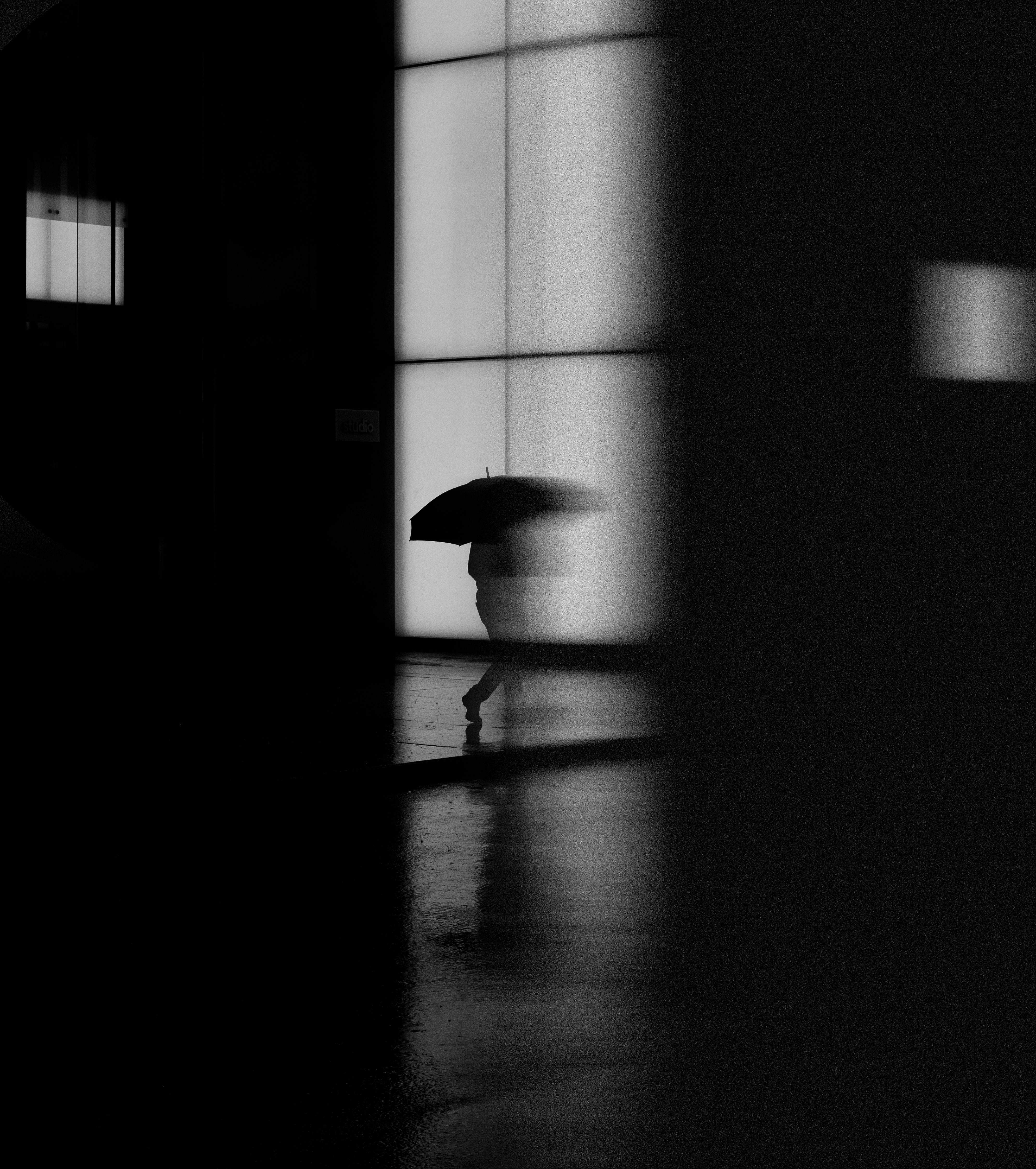 Silhouette of a person with an umbrella walking in the rain on a city street, captured in black and white.
