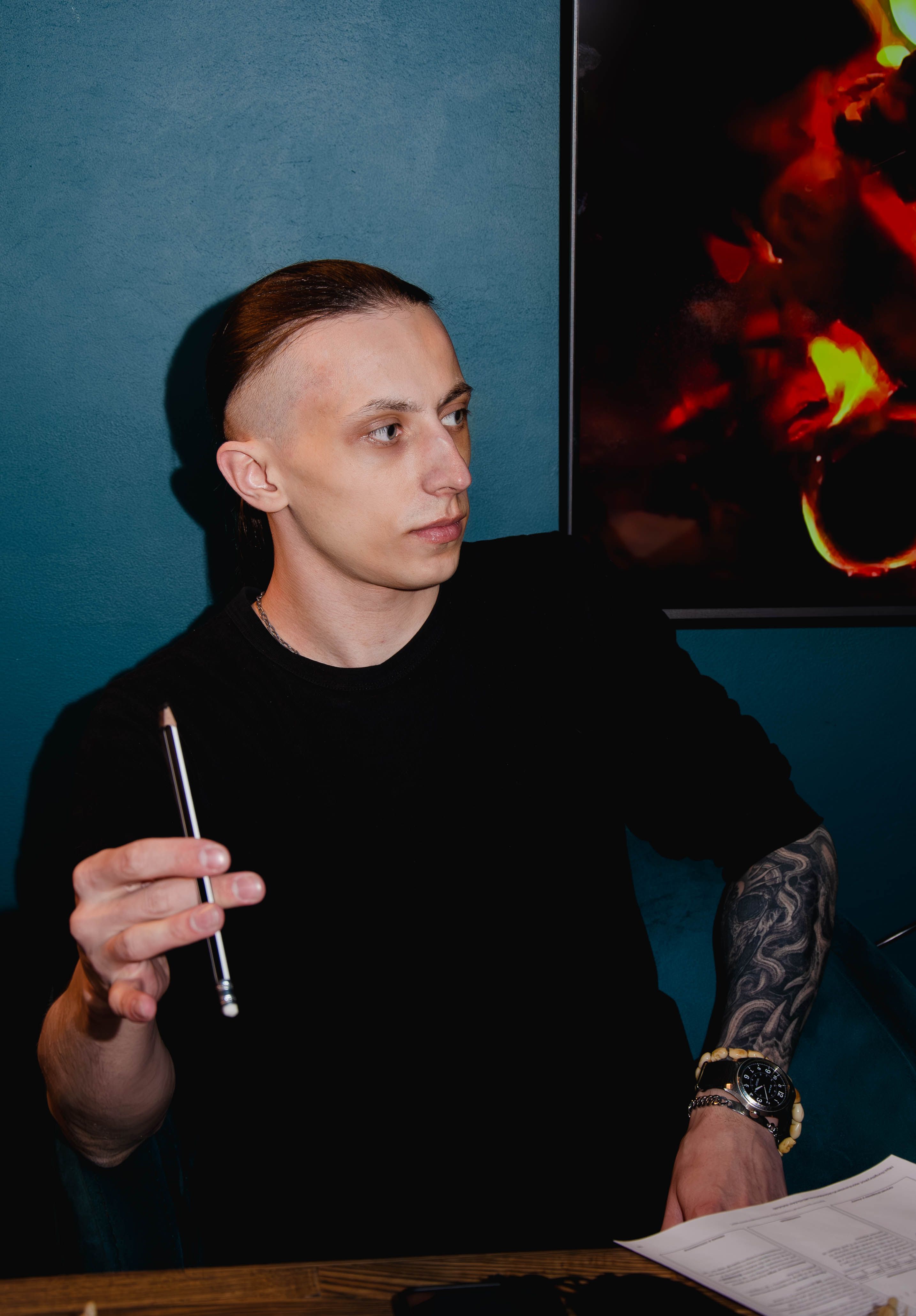 A young man with a tattooed arm holds a pencil, seated against a blue wall, showcasing focus and creativity.