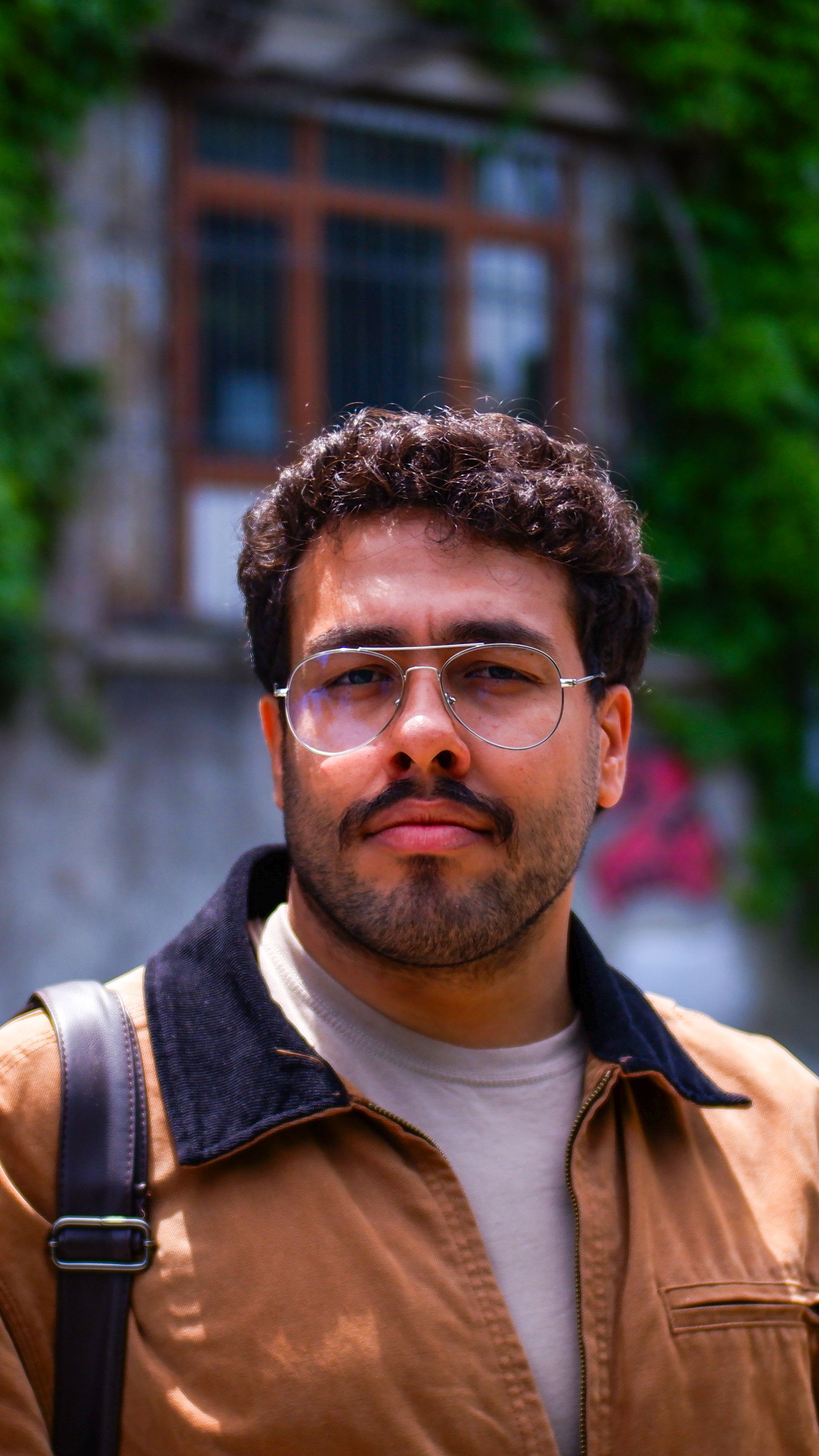 Casual portrait of a man wearing eyeglasses and a brown jacket outdoors with greenery in the background.