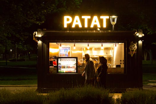 A couple walks past an illuminated 'Patat' stall at night in a park setting.