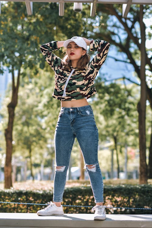 Photo of Woman in Camouflage Top and Denim Jeans Standing on a Stone Surface Posing with Her Hands on Her Head