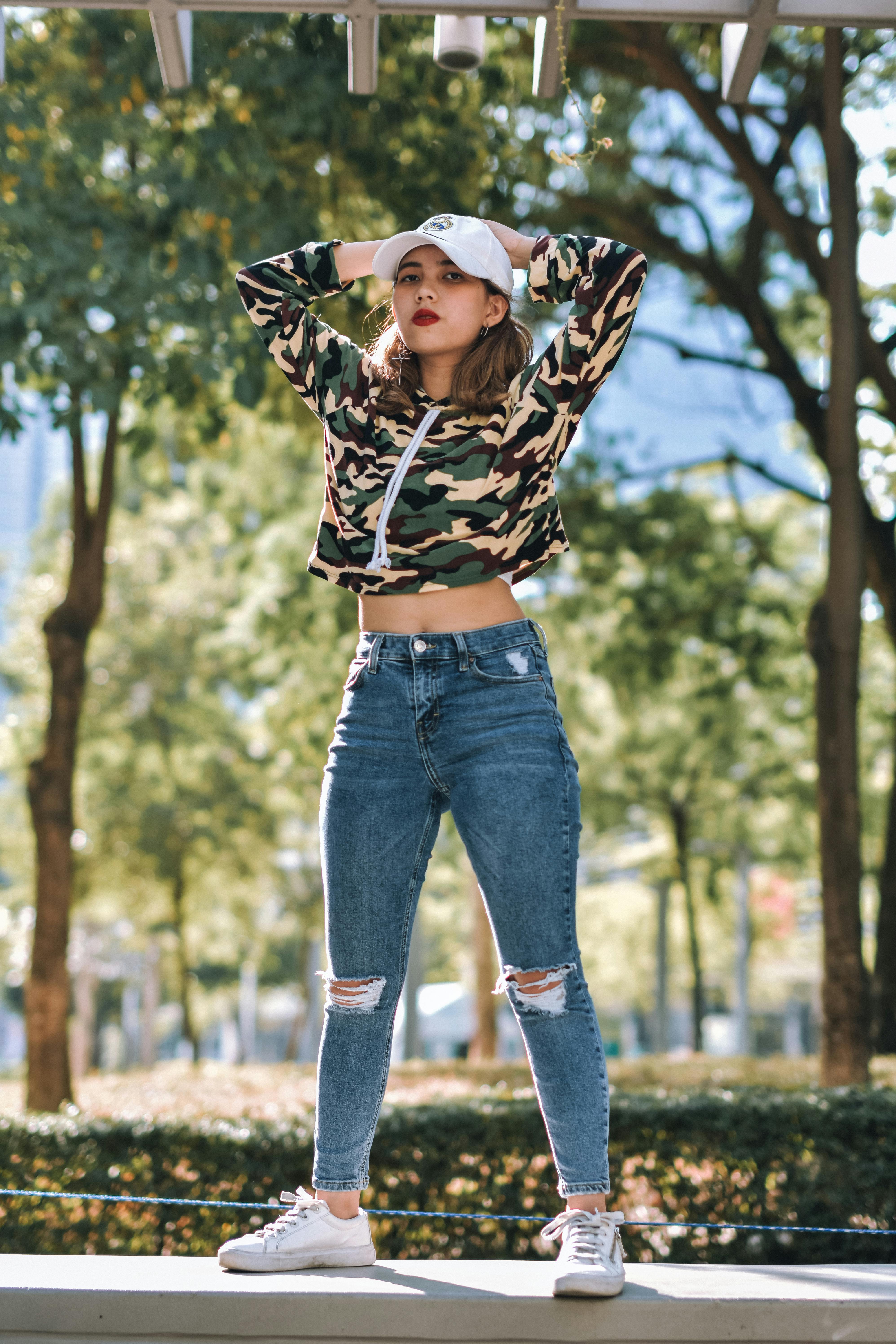 Photo of Woman in Camouflage Top and Denim Jeans Standing on a Stone Surface Posing Her Hands on Her Head Free Stock Photo