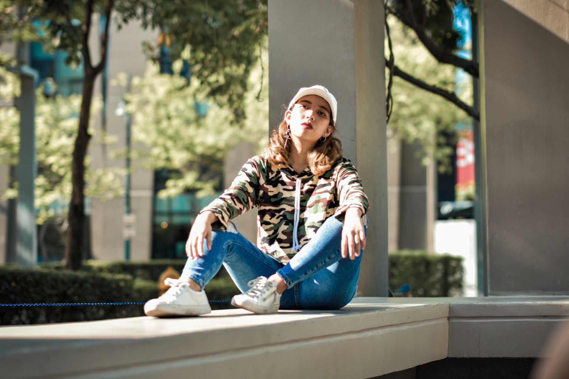 Femme assise en extérieur urbain portant une casquette blanche et un sweat camouflage