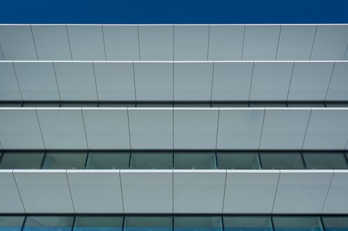 Free Low angle view of a modern building facade featuring geometric panels and glass windows under a clear blue sky. Stock Photo
