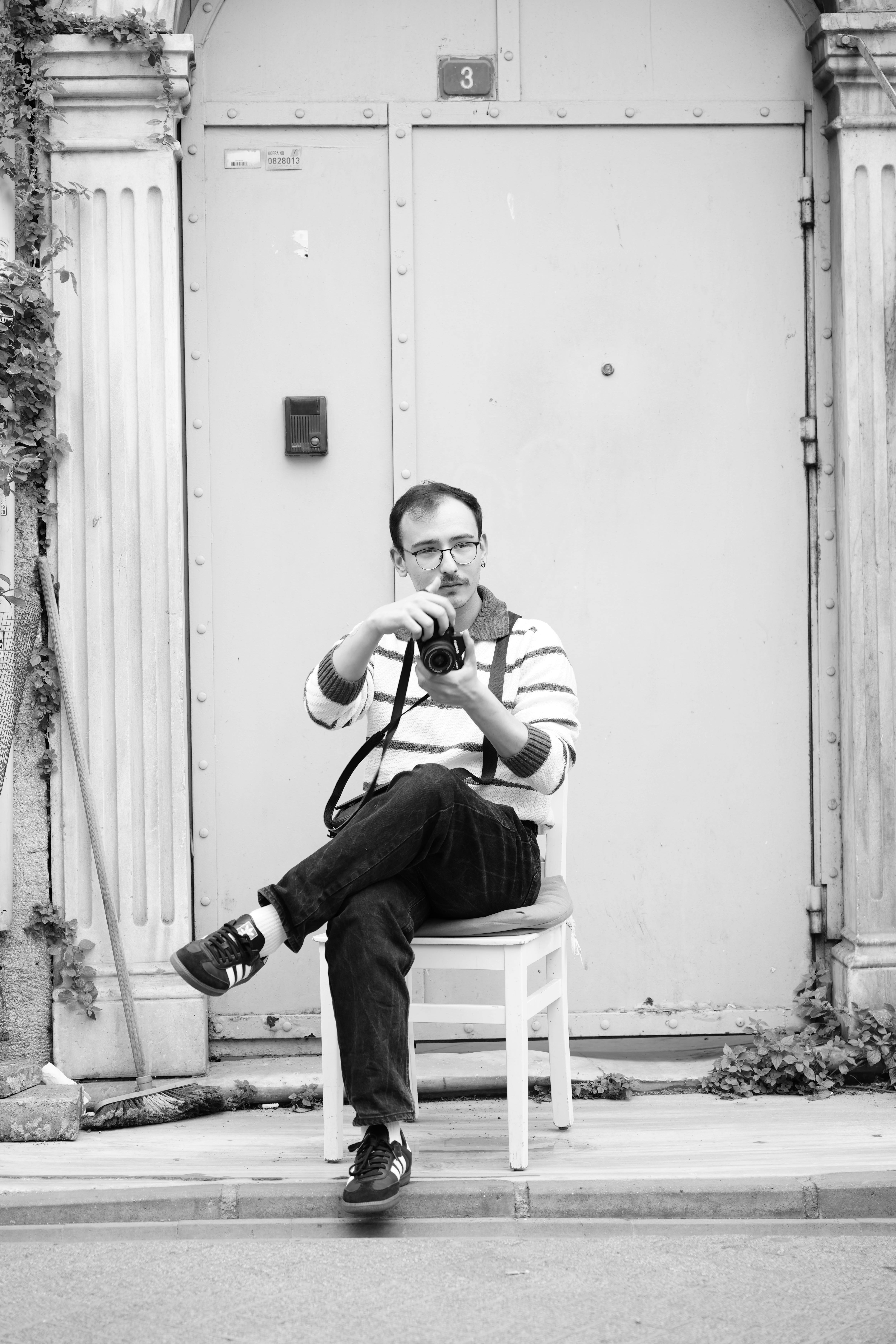 A man sits with a camera in an urban setting, embodying a modern fashion portrait.