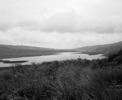 A tranquil black and white landscape of a lake and countryside under a cloudy sky.