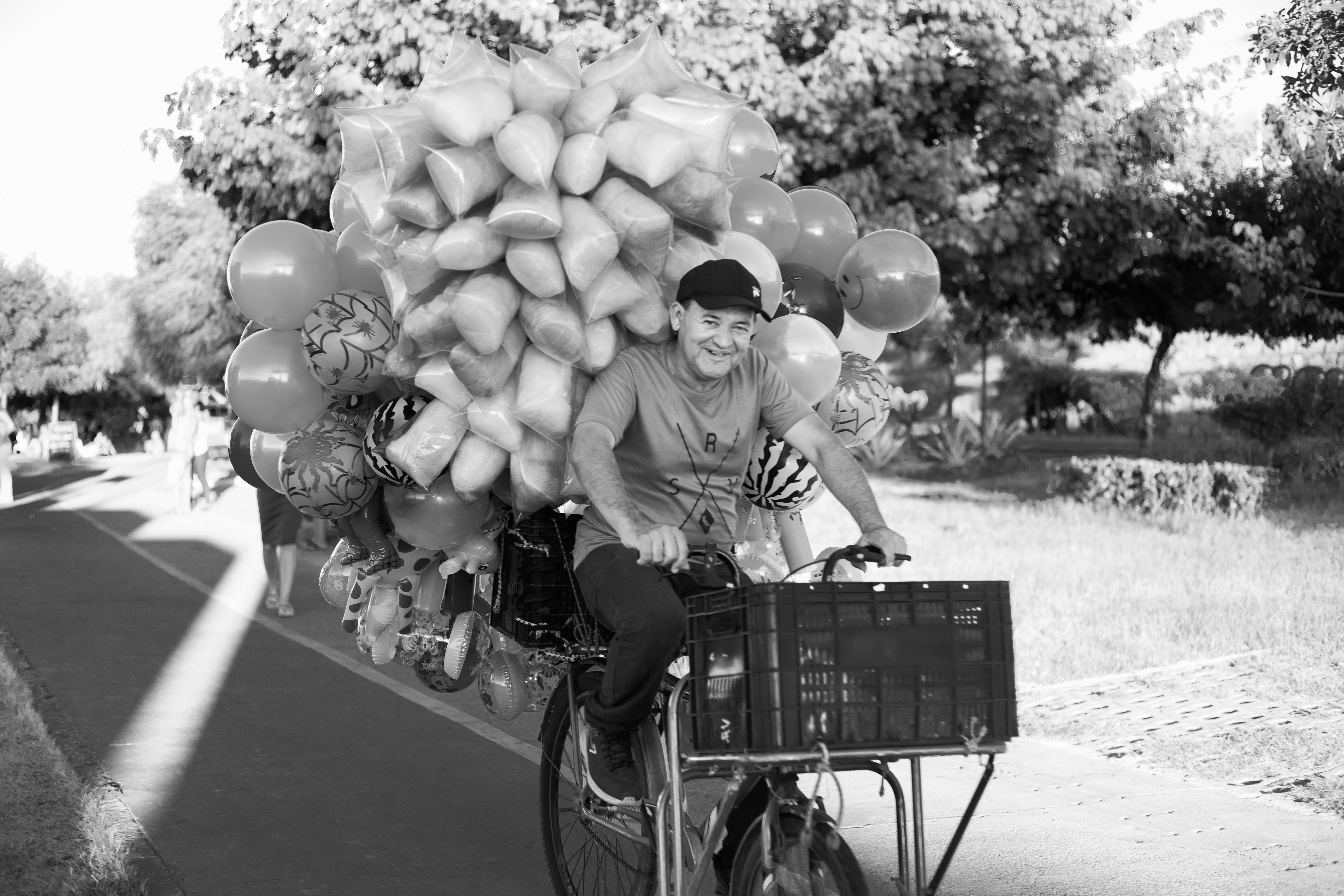 Smiling man rides bike loaded with balloons in an urban park setting.
