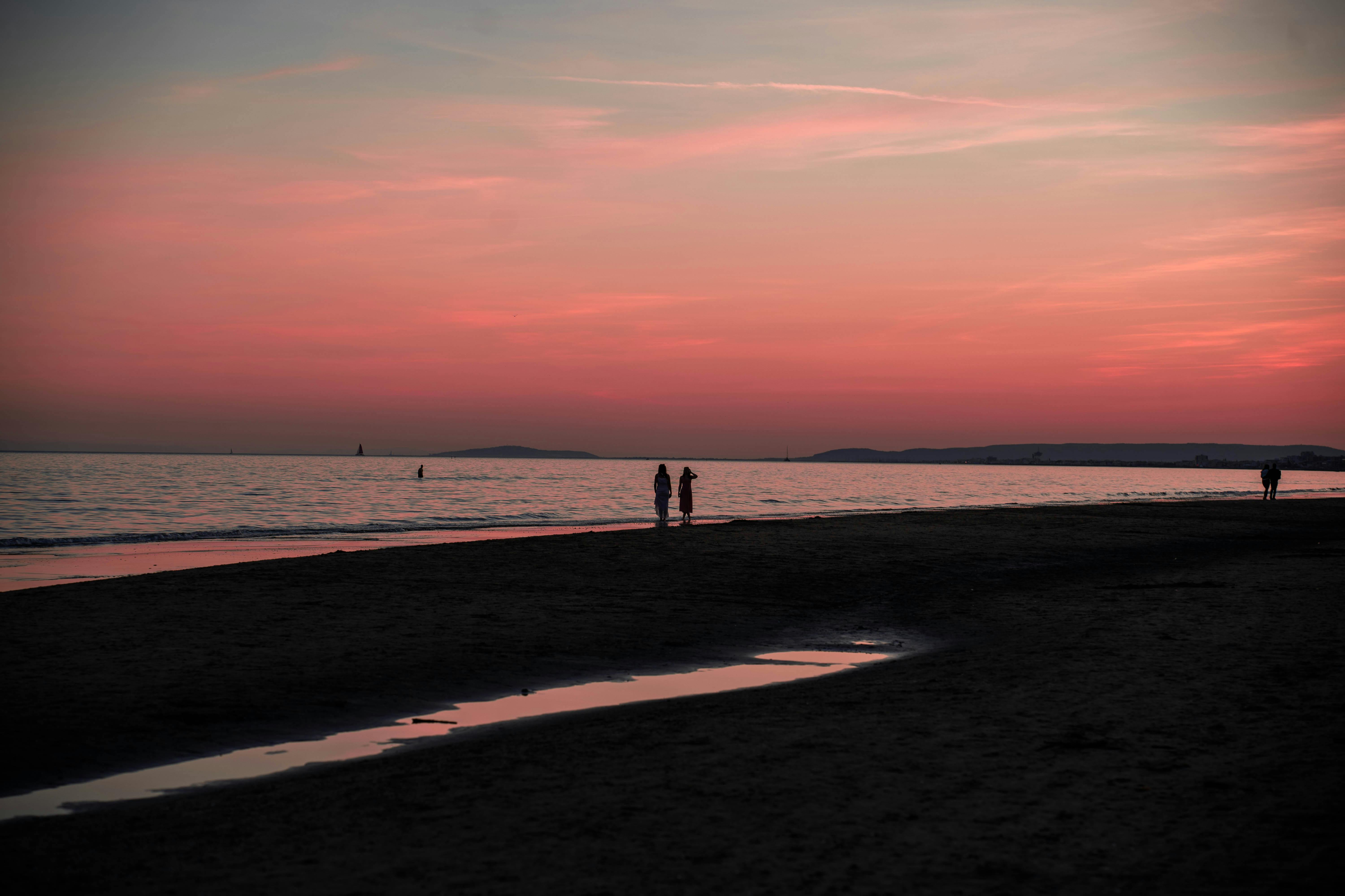Two people walk along the beach at sunset, silhouetted against a vibrant pink sky.