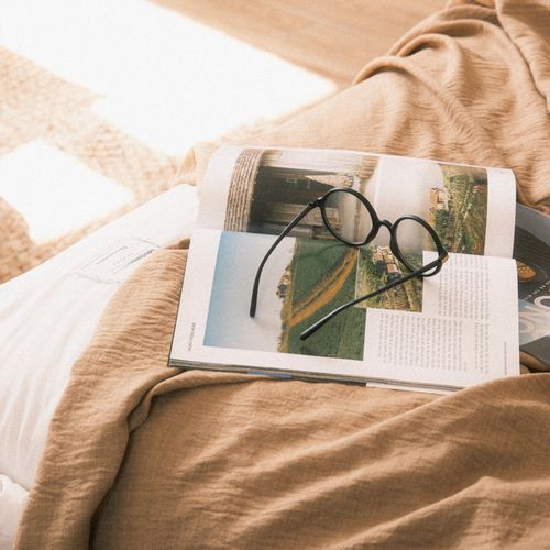 Free A pair of eyeglasses resting on an open book on a cozy bed, basked in gentle sunlight. Stock Photo
