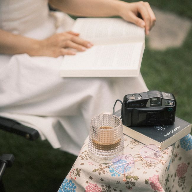 A woman seated outdoors reads a book with coffee and a camera nearby on a floral table.