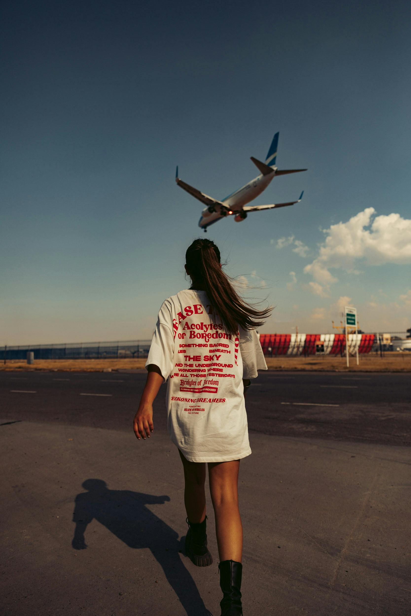 A woman in an oversized t-shirt walks on a road near an airport as an airplane flies overhead against a clear sky.