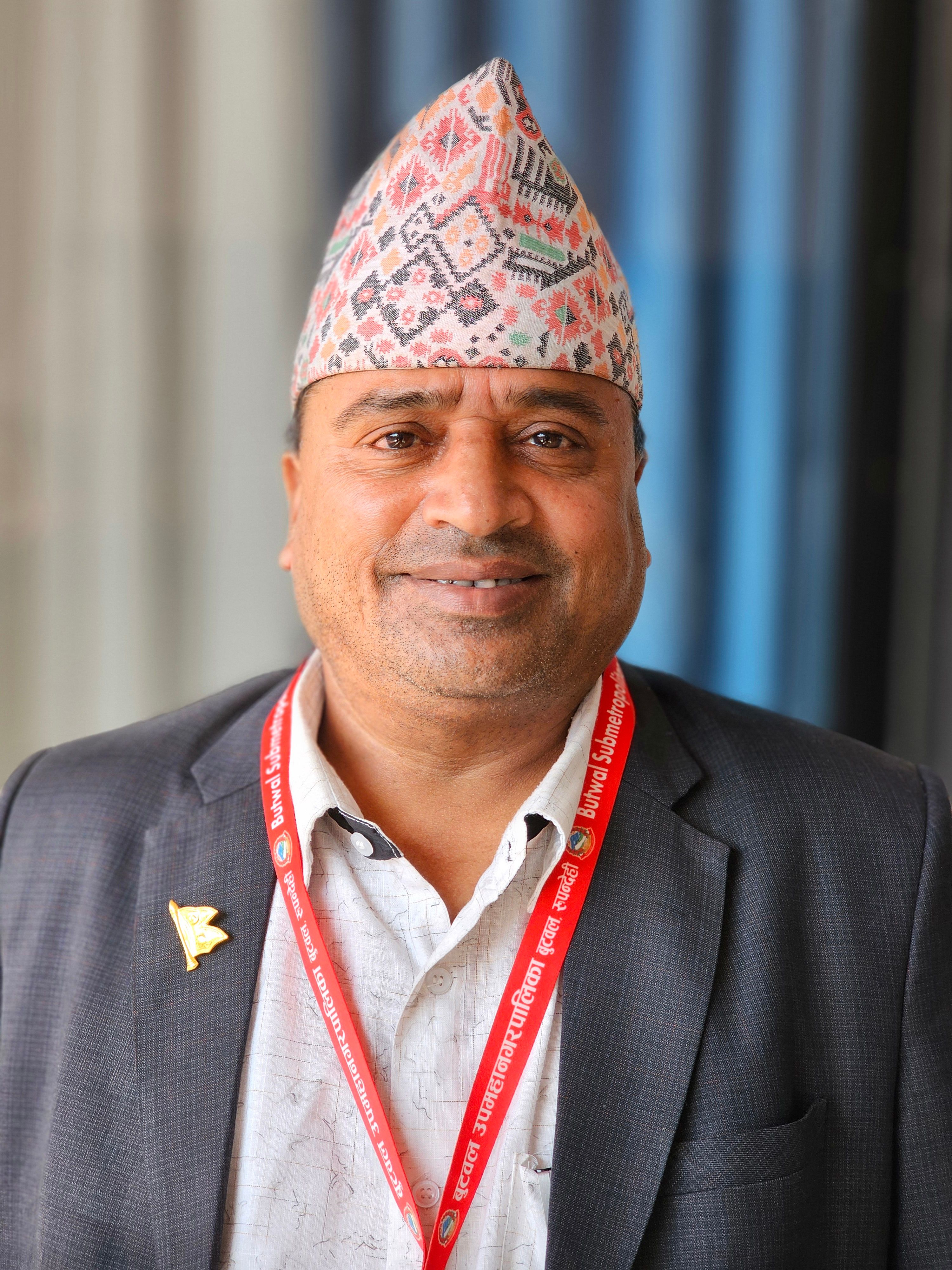 Smiling man wearing a traditional Nepali hat and a suit with a lanyard indoors.