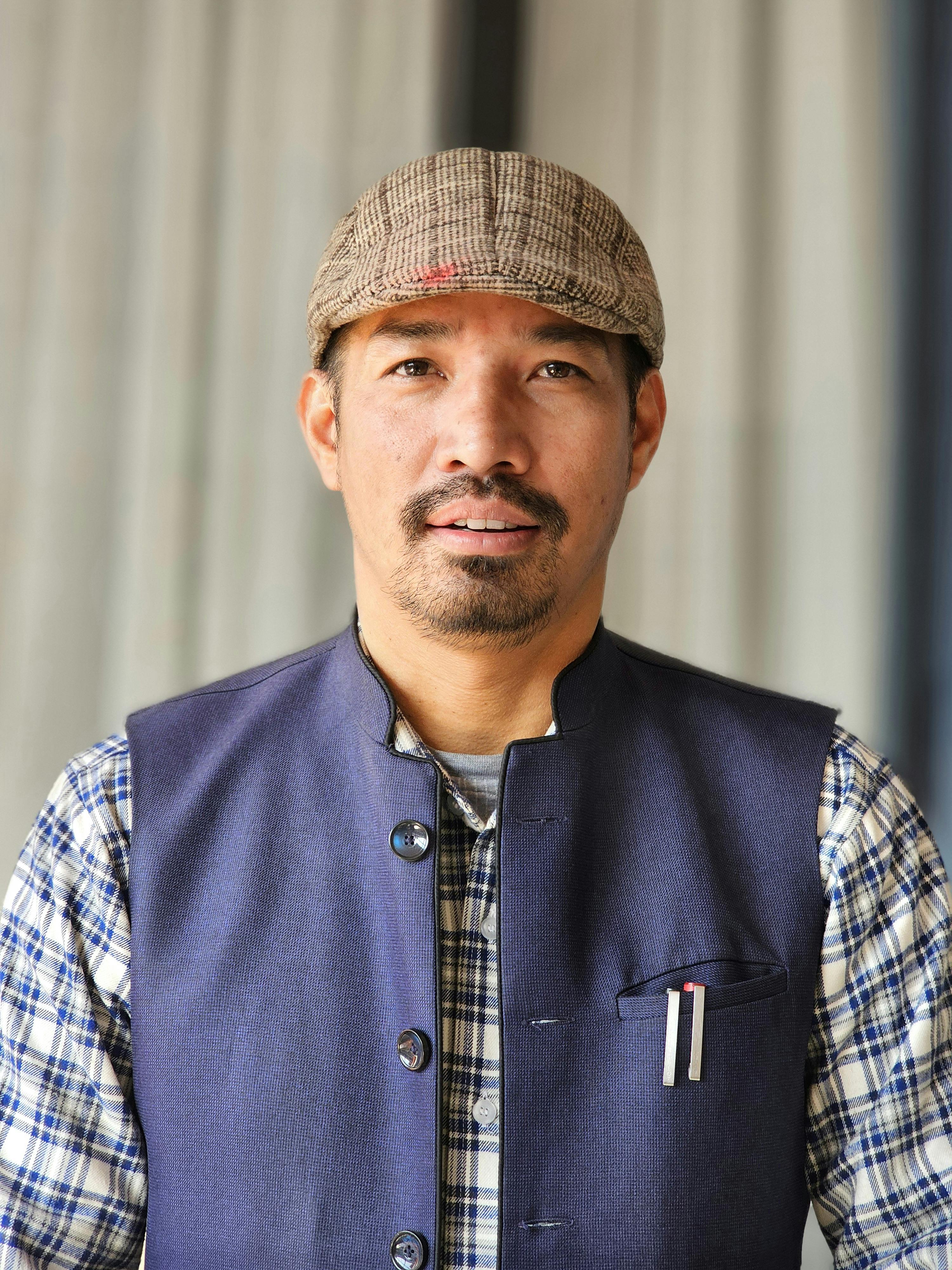A man wearing a flat cap and vest poses confidently indoors with a soft background.