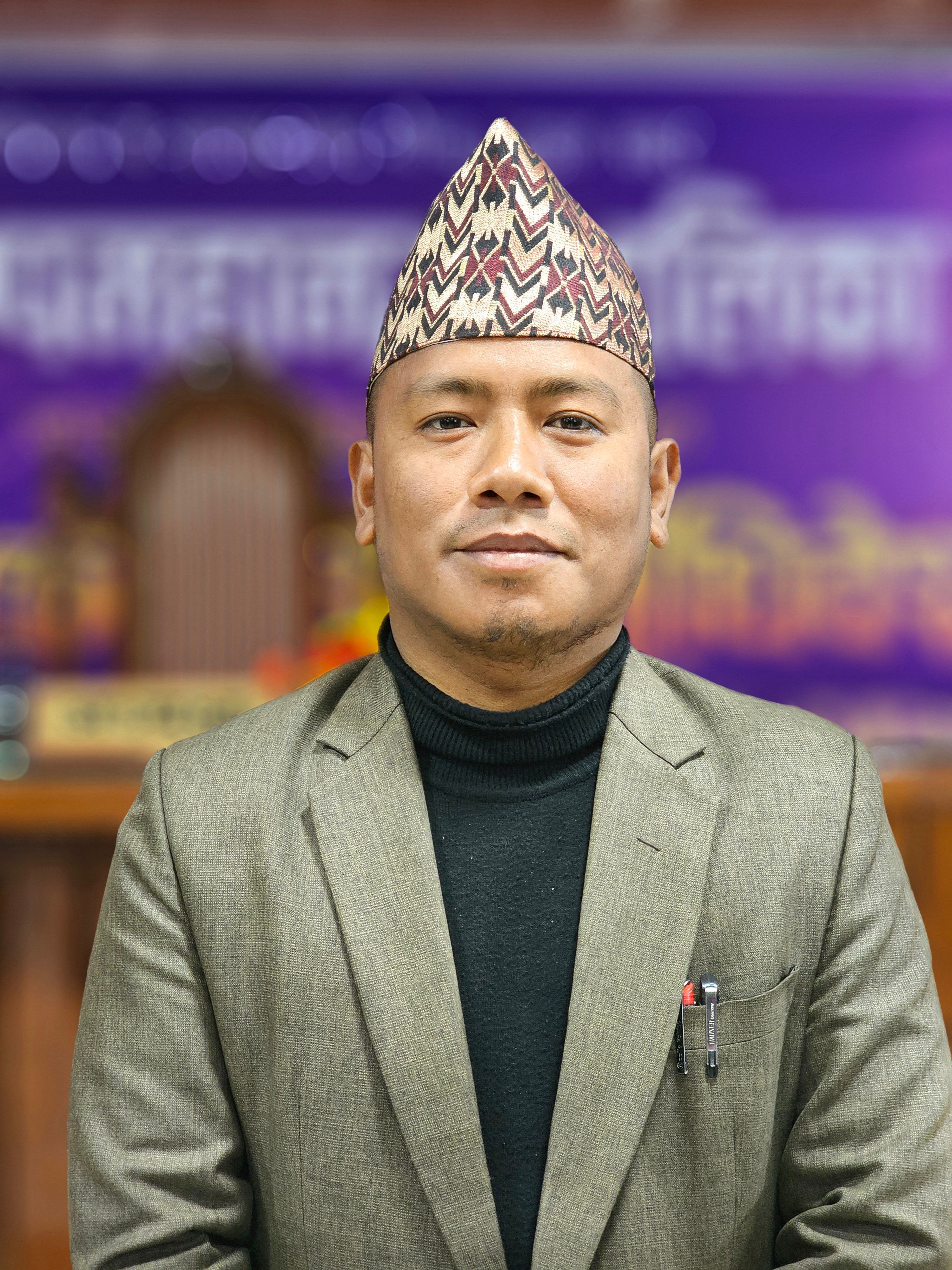 Portrait of a Nepali man wearing a Dhaka topi and suit, standing indoors.