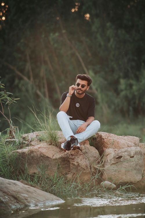 Free A young man wearing sunglasses sitting on rocks by a creek, enjoying a peaceful outdoor setting. Stock Photo