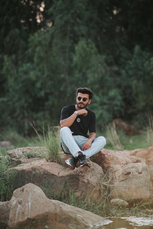 Free A casually dressed man sitting on rocks outdoors wearing sunglasses, t-shirt, and jeans. Stock Photo