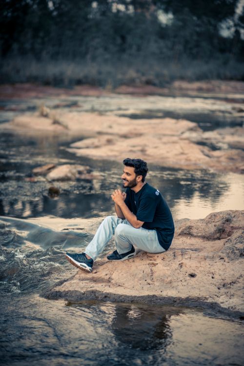 Free Man with beard and mustache sitting on rocks by the river, enjoying the peaceful outdoors. Stock Photo
