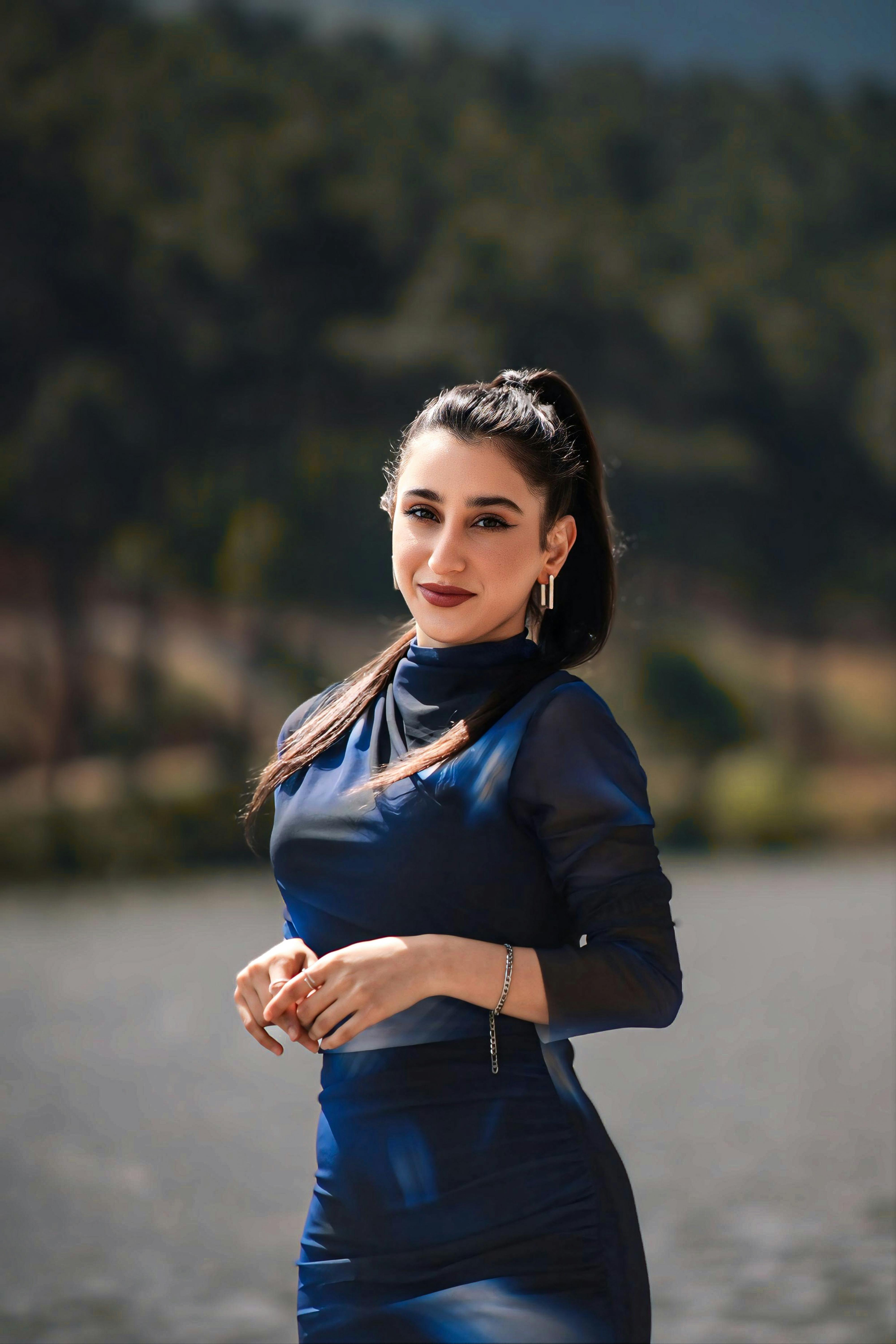 Stylish woman in a blue dress smiling by a serene lake under clear skies.