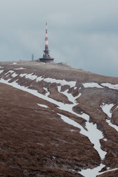 Telecommunication tower on Bucegi Mountain's snow-patched peak in Romania, overlooking a vast landscape.