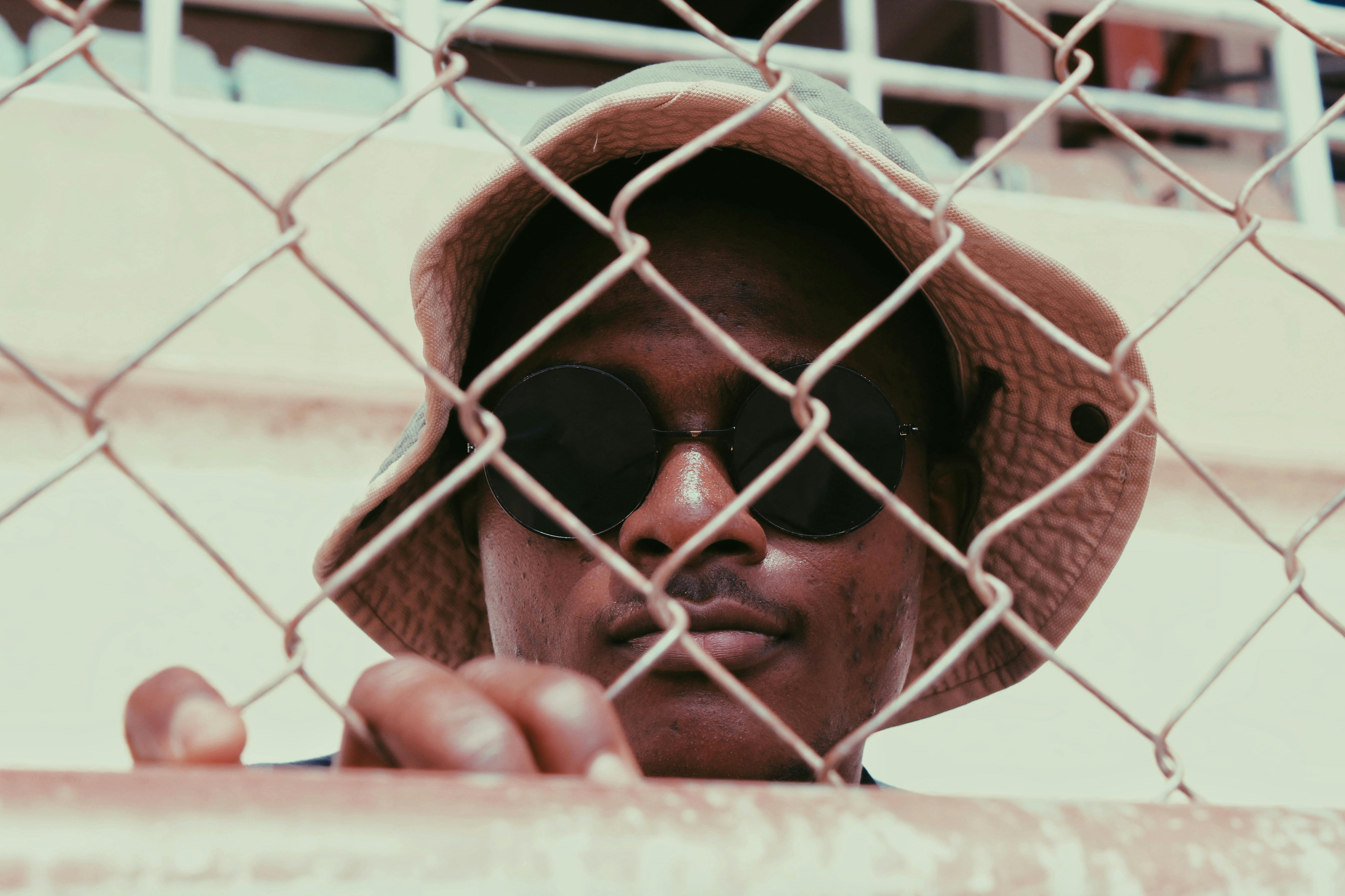 Photo of a man wearing black sunglasses standing behind a chain link fence · Free Stock Photo