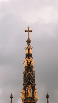 Close-up of a Gothic-style monument in London with a decorative cross and golden statues against a cloudy sky.