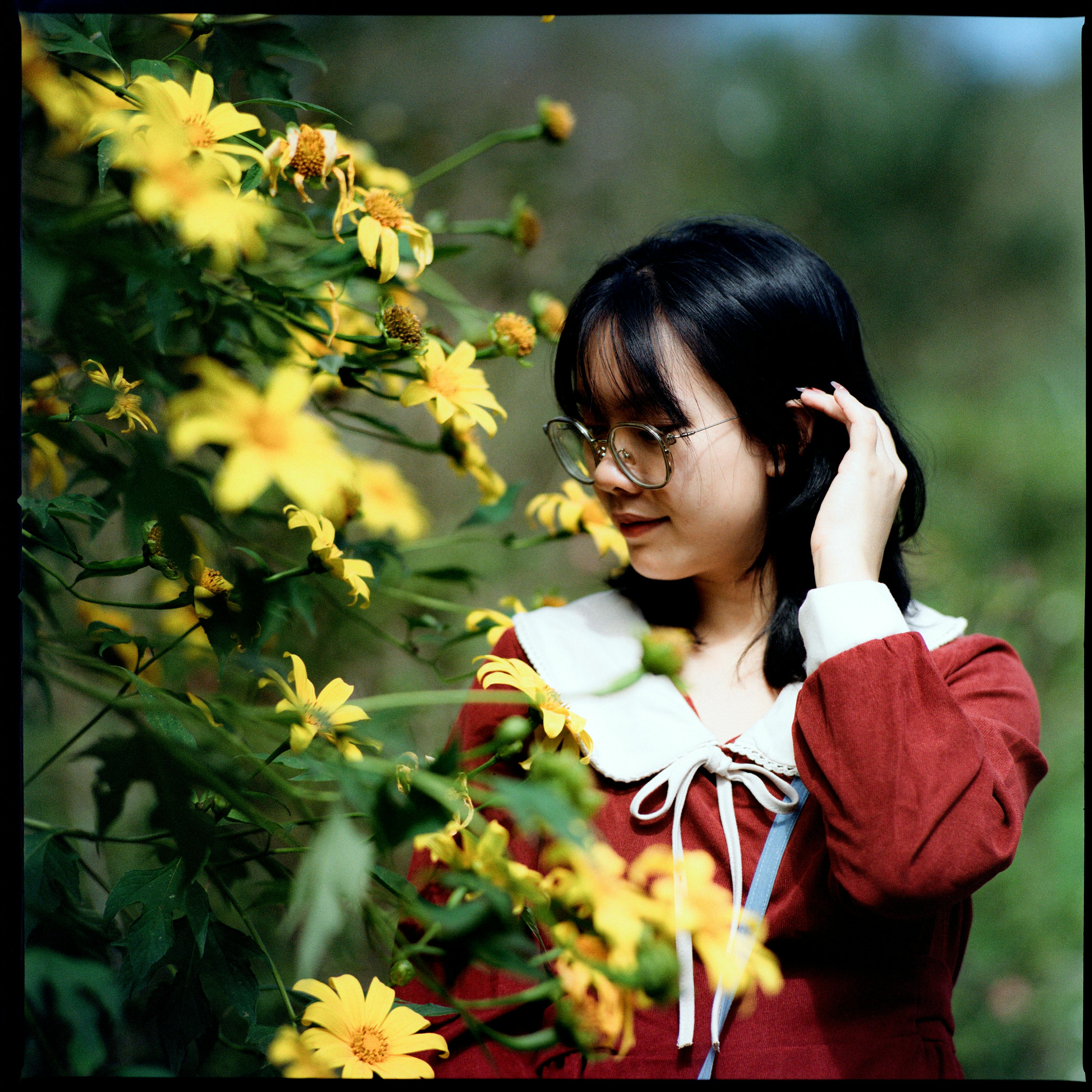 Young woman in a red dress with eyeglasses among vibrant yellow flowers in a garden setting.