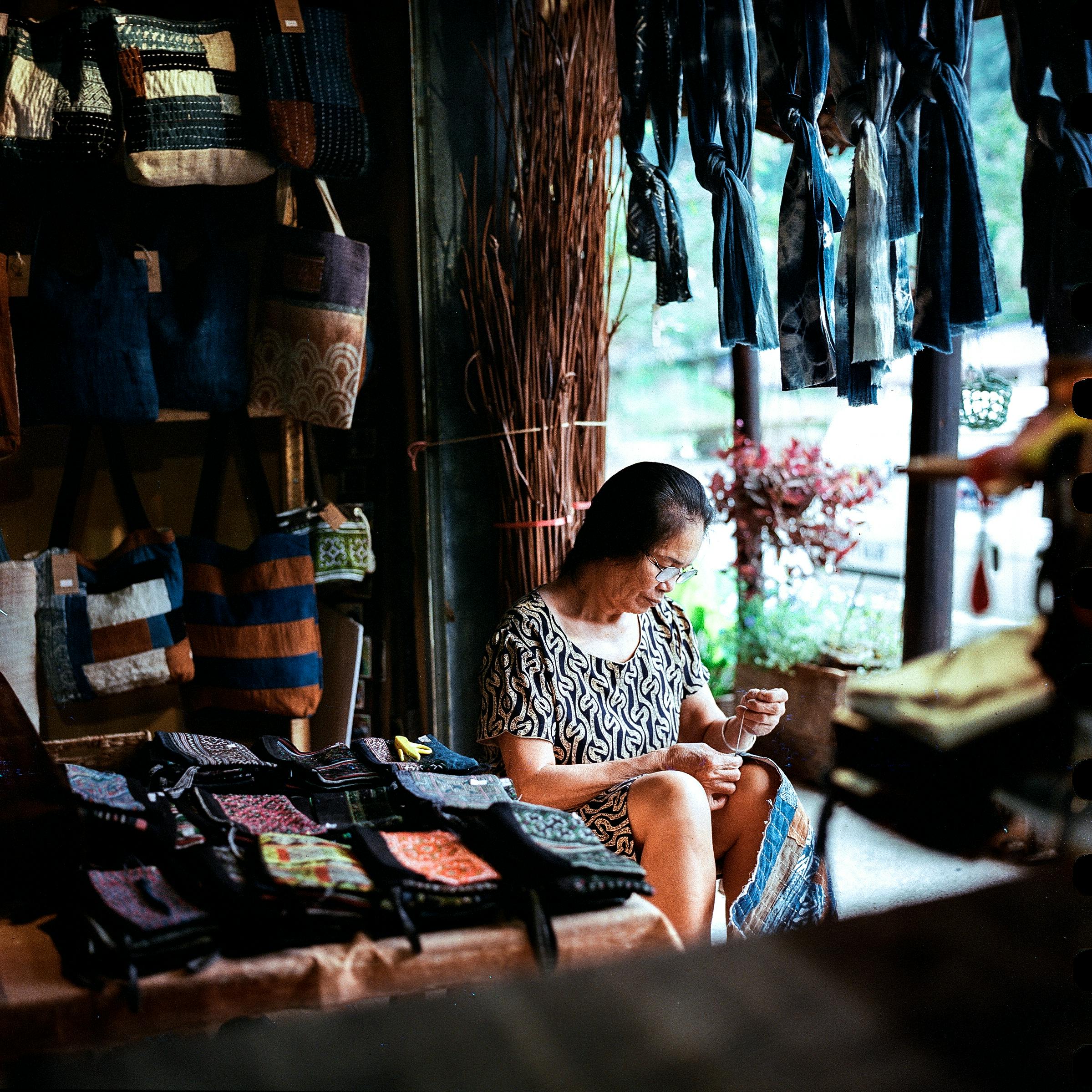 An elderly Asian woman intricately sewing in a cozy artisan store setting.