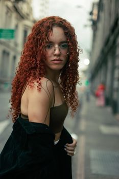 Stylish young woman with glasses and curly red hair poses on an urban city street.